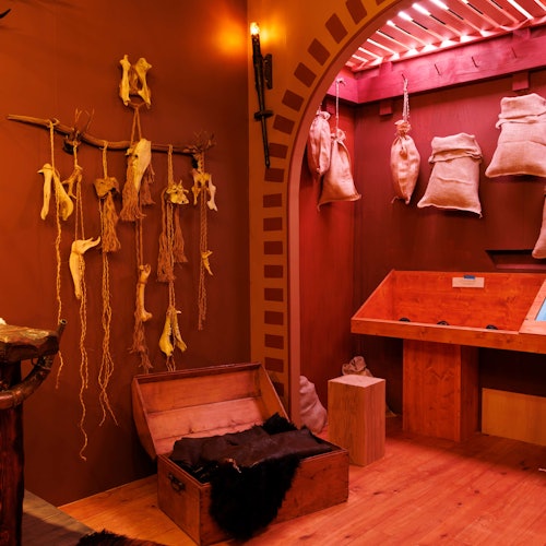 Room with wooden chests, hanging dried plants, and burlap sacks. Warm lighting and rustic decor create a cozy ambiance.