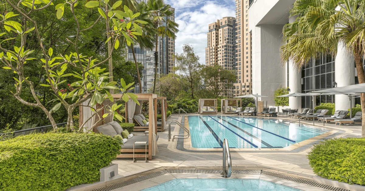 An outdoor swimming pool area surrounded by greenery and tall city buildings, featuring lounge chairs and a jacuzzi