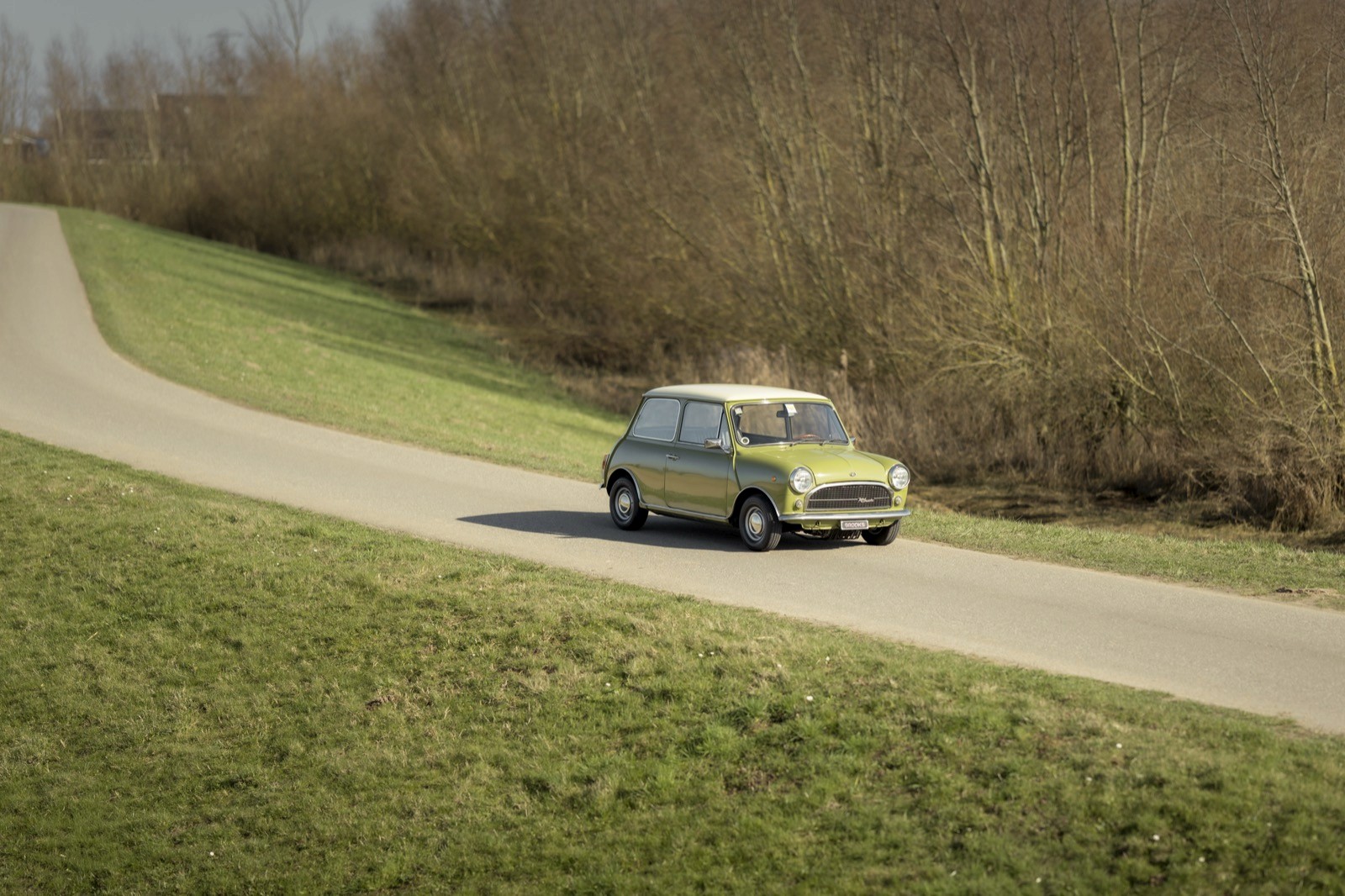 A green car drives on a winding road surrounded by grass and bare trees under a clear sky.