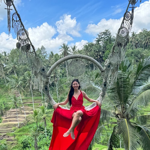 風の遊園地にある美しい緑の棚田の上で、レッドロングドレスを着た女性がドリームキャッチャーブランコに乗っています。