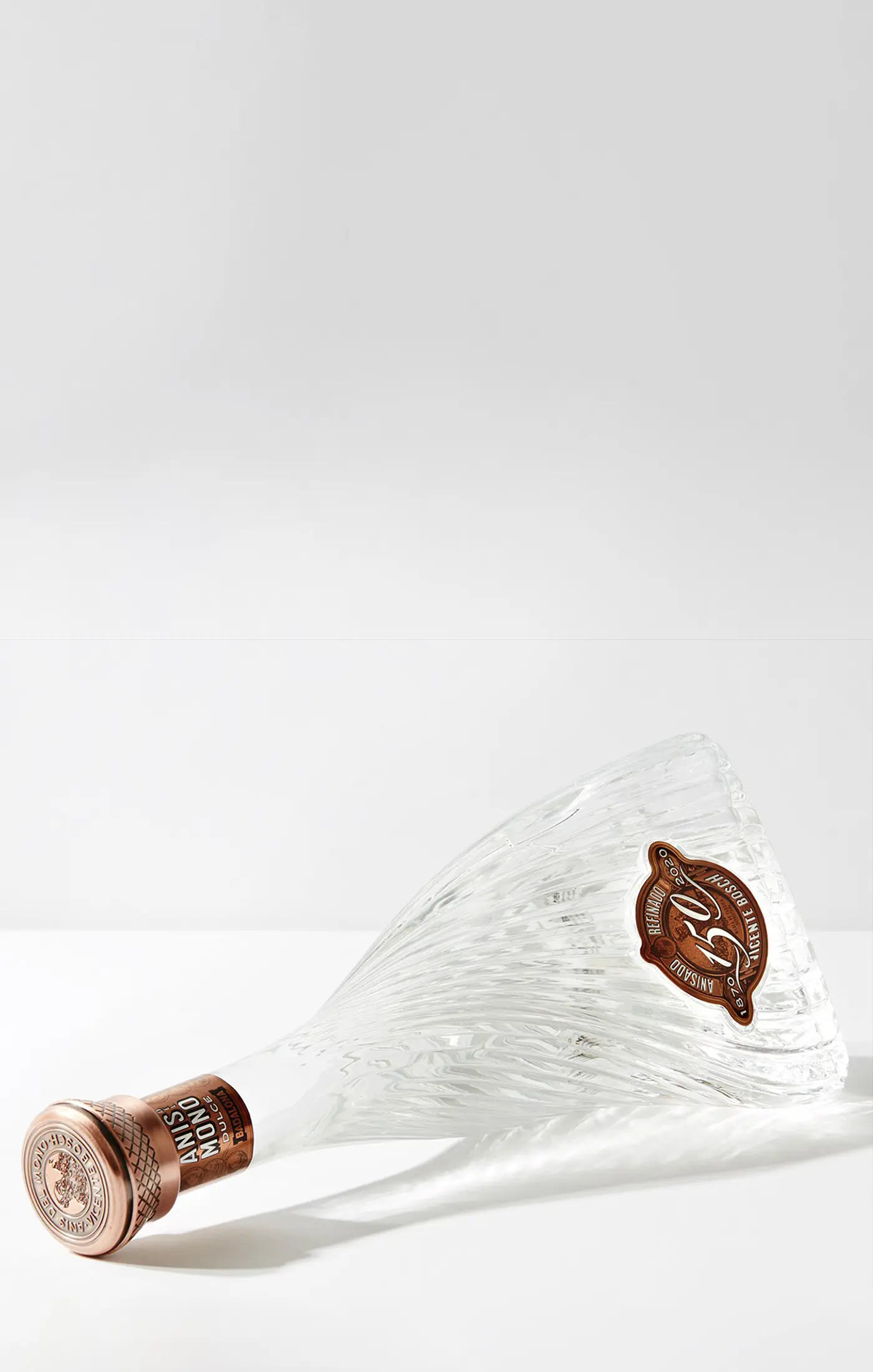Elegant glass bottle with textured swirls lies on its side. Copper-toned cap and label read "ANIS NOVO". Minimalist and sophisticated ambiance.
