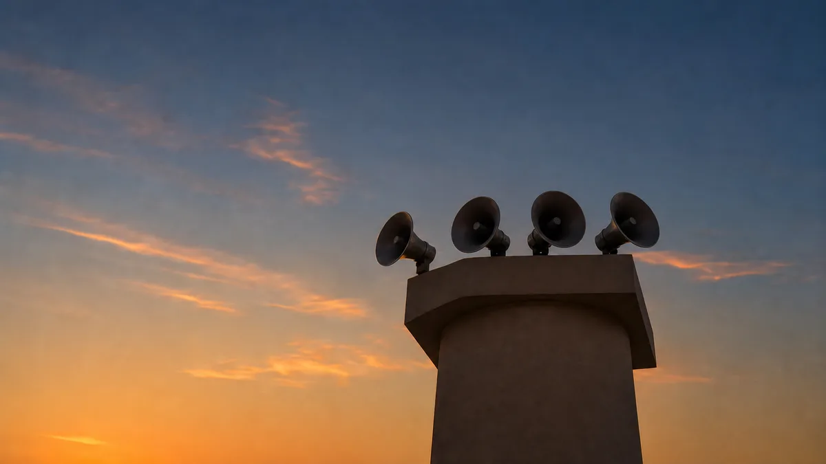 Four plain loudspeaker horns on a minaret silhouetted against an amber-blue dusk sky
