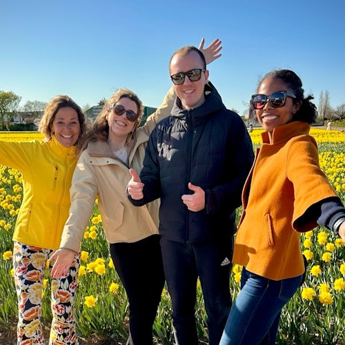 Apat na tao ang nakangiti at nagpo-pose sa isang parang ng dilaw na mga bulaklak sa ilalim ng maliwanag na asul na kalangitan.