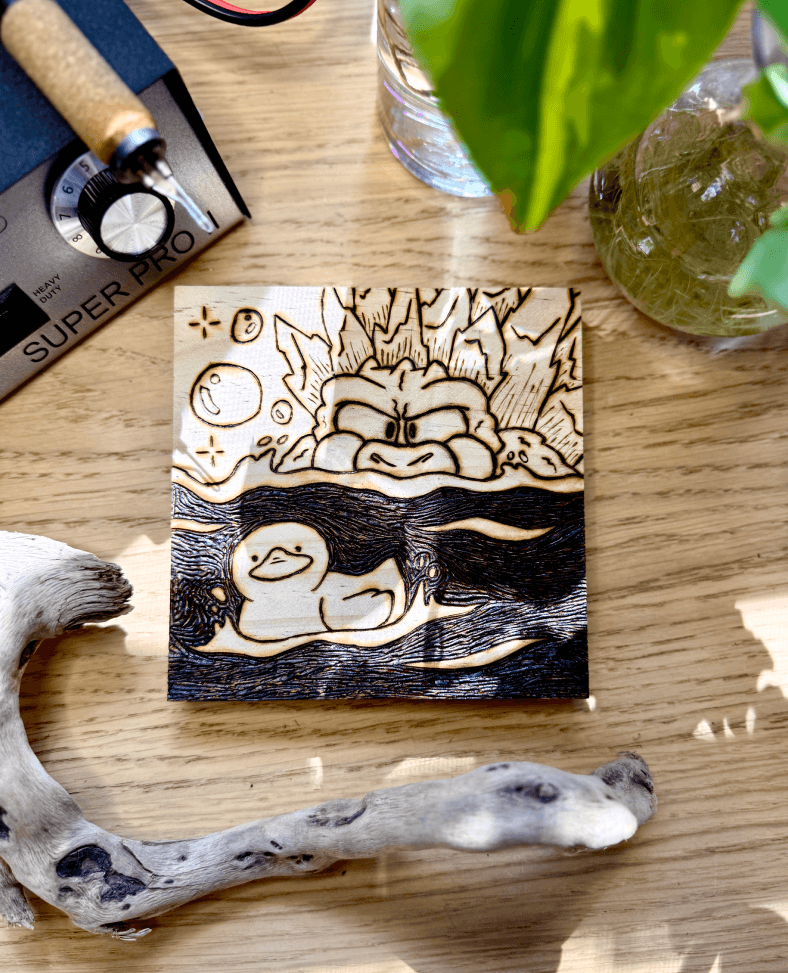 A wood burned decorative plaque depicting Godzilla in the water stalking down a rubber duck. Bubbles are floating to his left. This is a reimagined piece honoring Godzilla Minus Zero. The plaque is surrounded by plants, drift wood, and the colwood pro 2 wood burner.