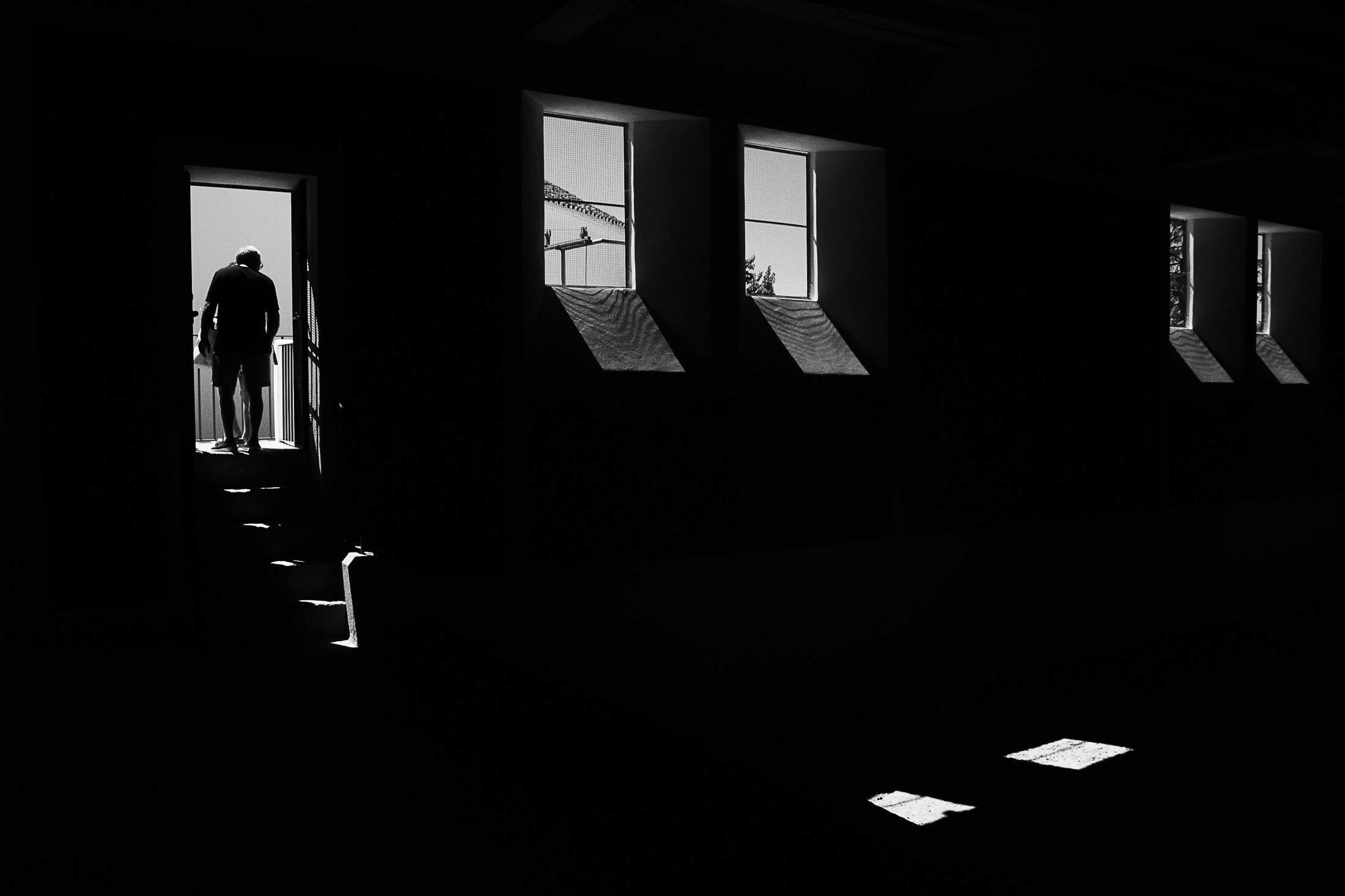 Black and white photograph of a man standing in a doorway illuminated by window light in a dark interior.