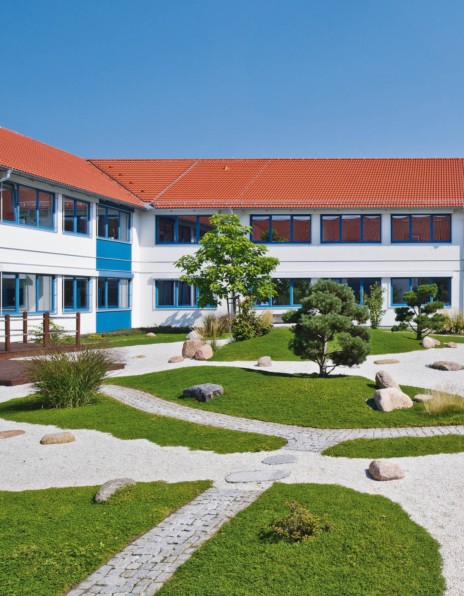 Moderne weiße Gebäudestruktur mit roten Ziegeldächern und blau gerahmten Fenstern inmitten eines gestalteten Gartens.