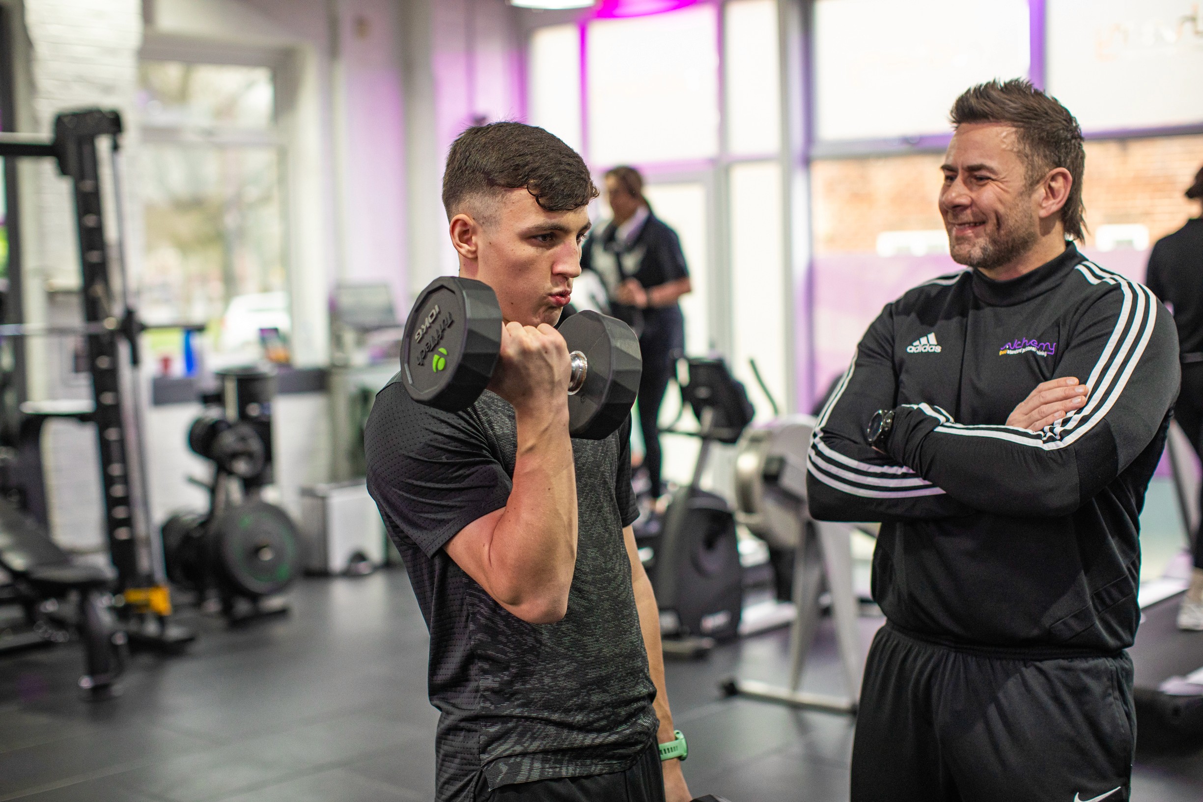 A young man is doing a dumbbell curl in a gym while a personal coach watches and smiles. There are other gym equipment and machines in the background.
