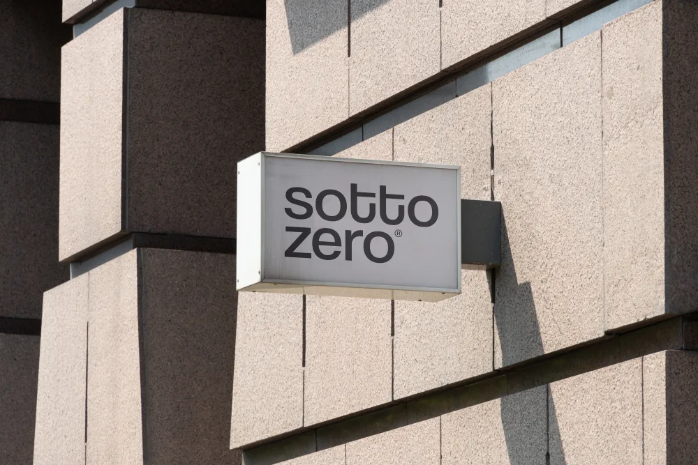Outdoor signage mockup mounted on a beige textured concrete wall. The rectangular sign displays minimalist black-and-white branding with bold typography. Sunlit urban setting.