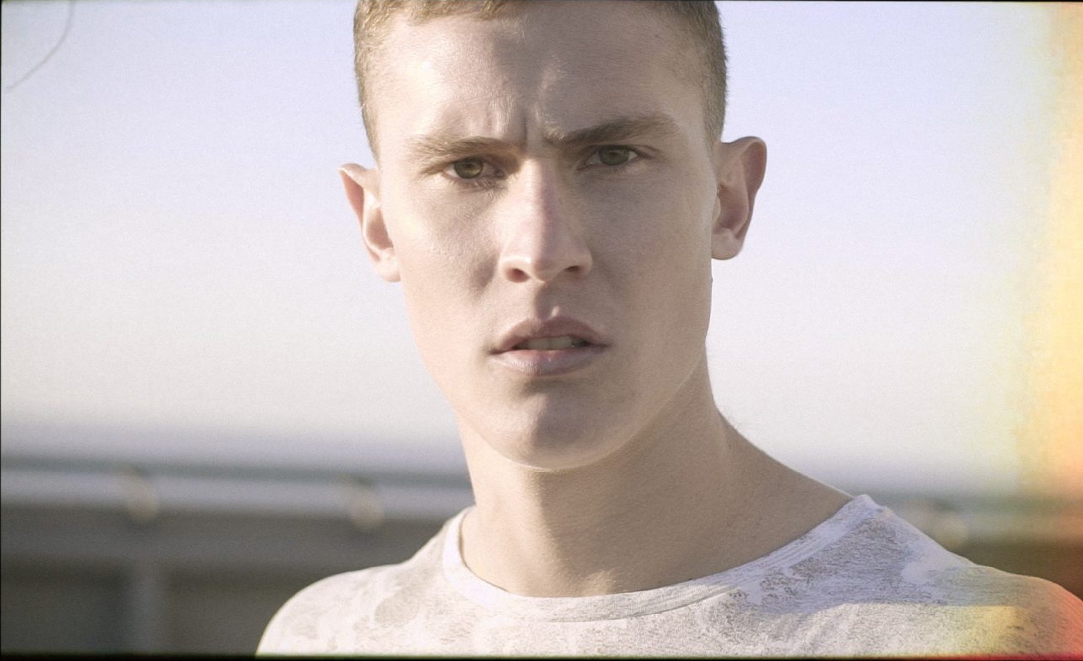 A young man stands outdoors, looking directly at the camera with a serious expression. He wears a light-colored shirt and is set against a blurred background.