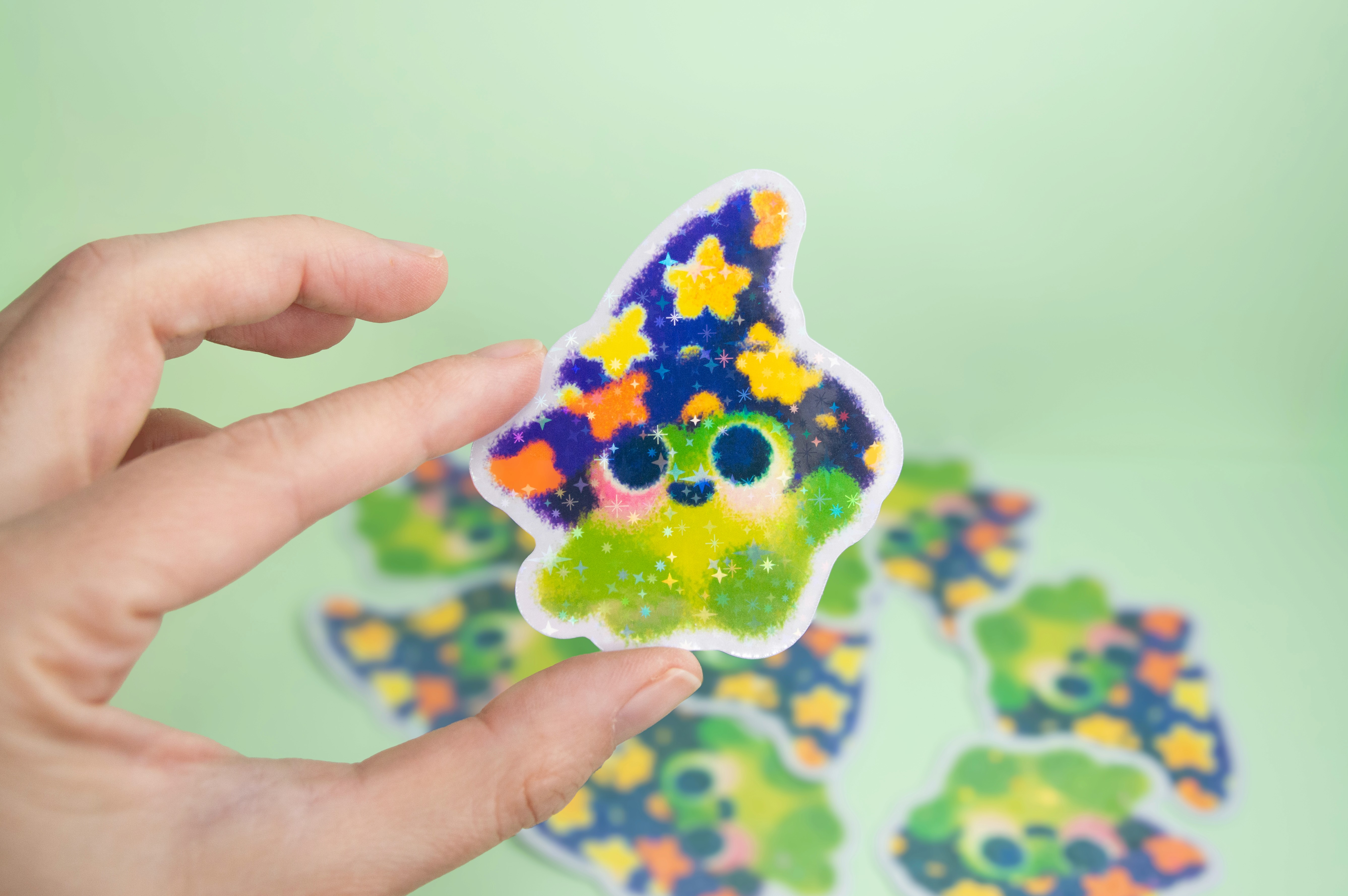 A hand holding a sparkling sticker featuring a cute frog illustration. The frog is wearing a starry wizard hat.