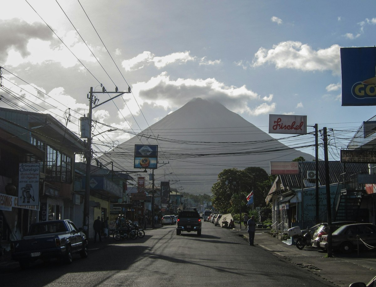 Arenal Volcano rising beyond a street of shops, power lines and parked cars in La Fortuna