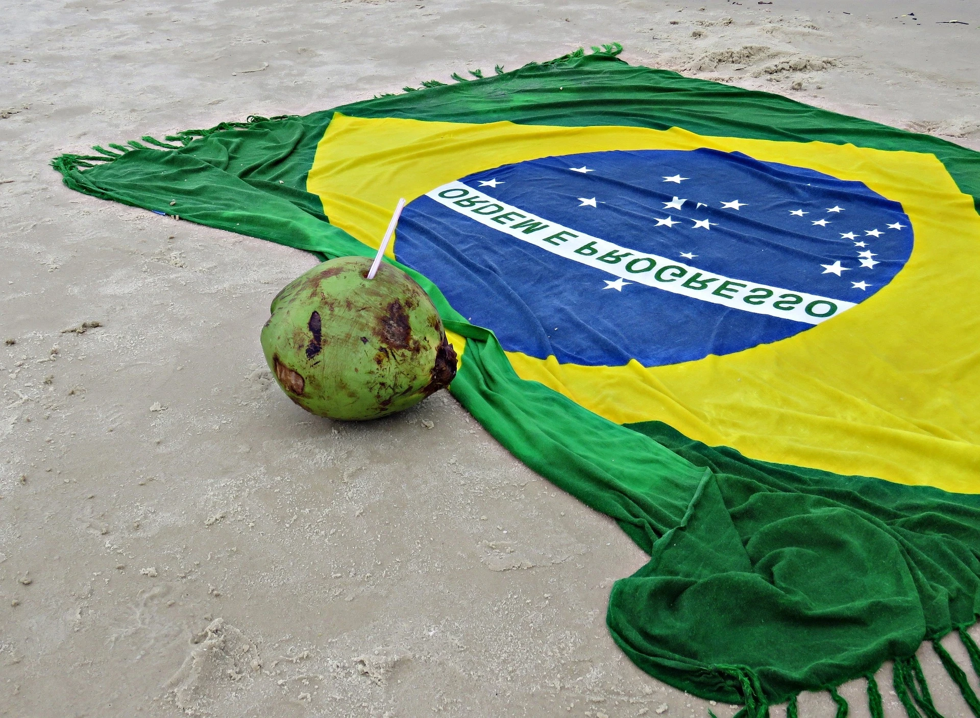 Toalla con la bandera de Brasil y un coco con sorbete sobre la arena, ideal para turistas que buscan cómo pagar con Pix en Brasil mientras disfrutan de la playa.