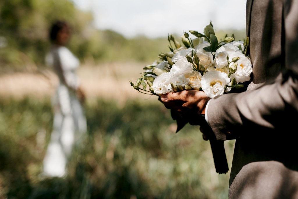 Groom holding white rose bouquet with bride in the background