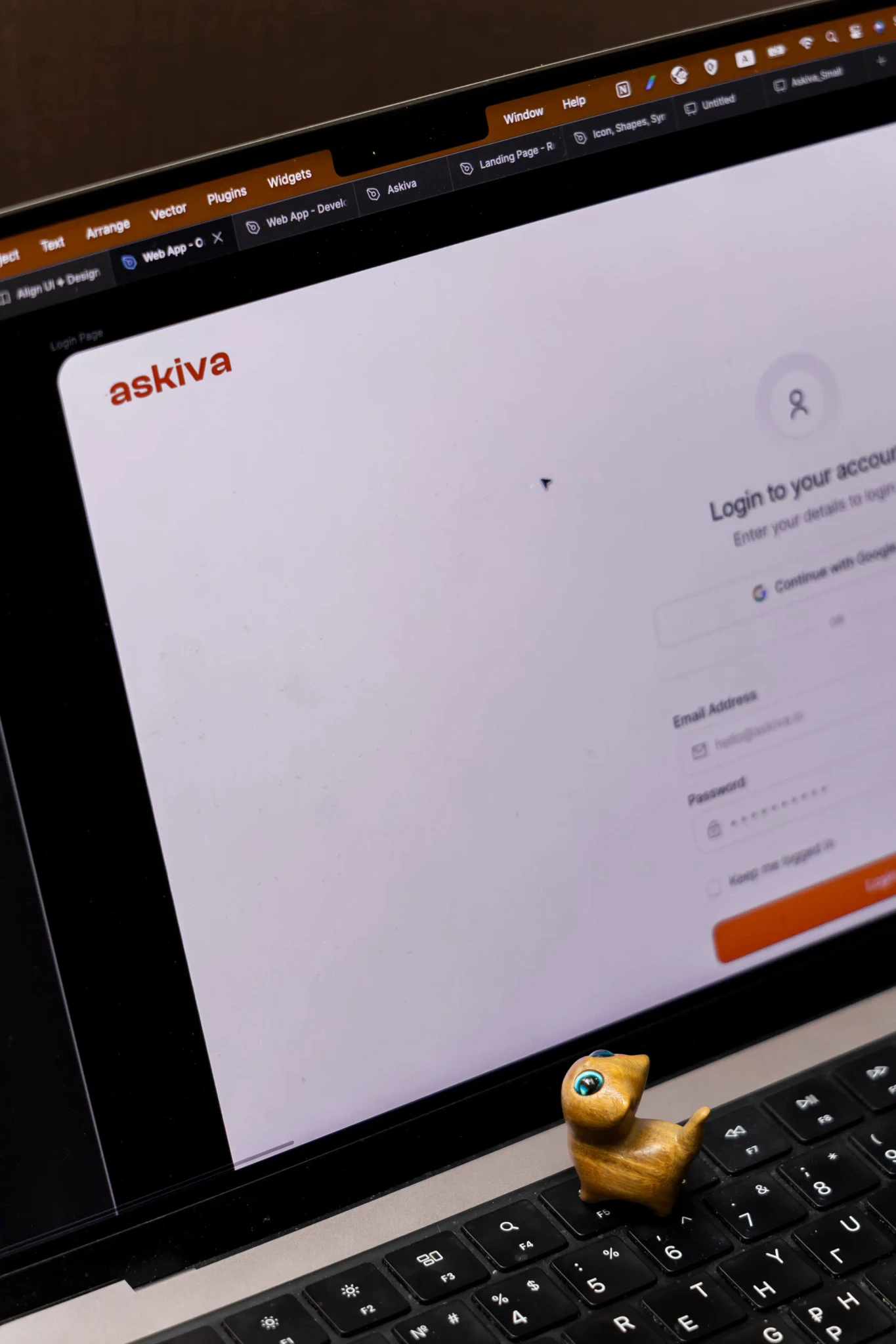 Close-up of a laptop showing the Askiva login screen, with a small figurine on the keyboard