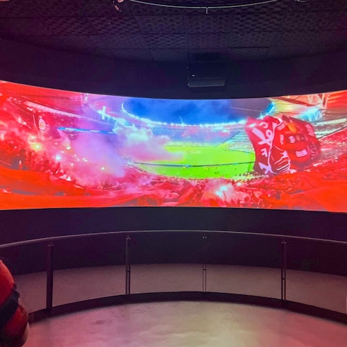 在一个黑暗的房间里，从栏杆障碍物后观看，一块大型弧形屏幕正在展示一个色彩缤纷、观众满满的体育场，正进行着一场热闹的活动。