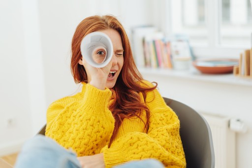 A person in a bright yellow sweater playfully holds a disc over their eye, sitting in a cozy indoor setting.