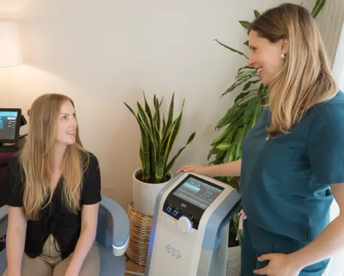 Female healthcare professional in teal scrubs explaining a pelvic floor therapy procedure to a seated patient beside an Emsella treatment chair.