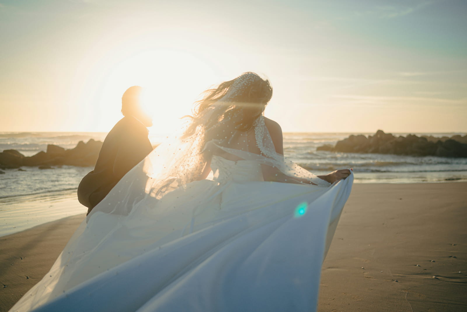Bride's dress and hair flowing in the ocean breeze at sunset on a rocky shoreline with the groom silhouetted by golden light