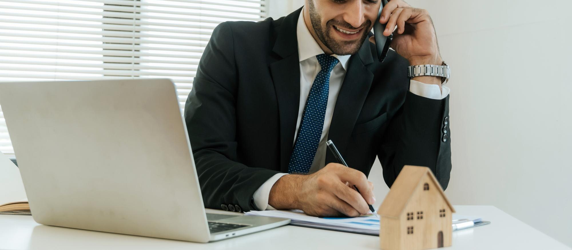 Makelaar aan bureau met telefoon en laptop, houten huismodel op tafel — AI klantenservice voor makelaars