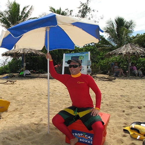 บุคคลสวมเสื้อแรชการ์ดสีแดงนั่งอยู่ใต้ร่มสีฟ้าขาวบนชายหาดที่เต็มไปด้วยทราย มีต้นปาล์มและผู้คนอยู่ในฉากหลัง