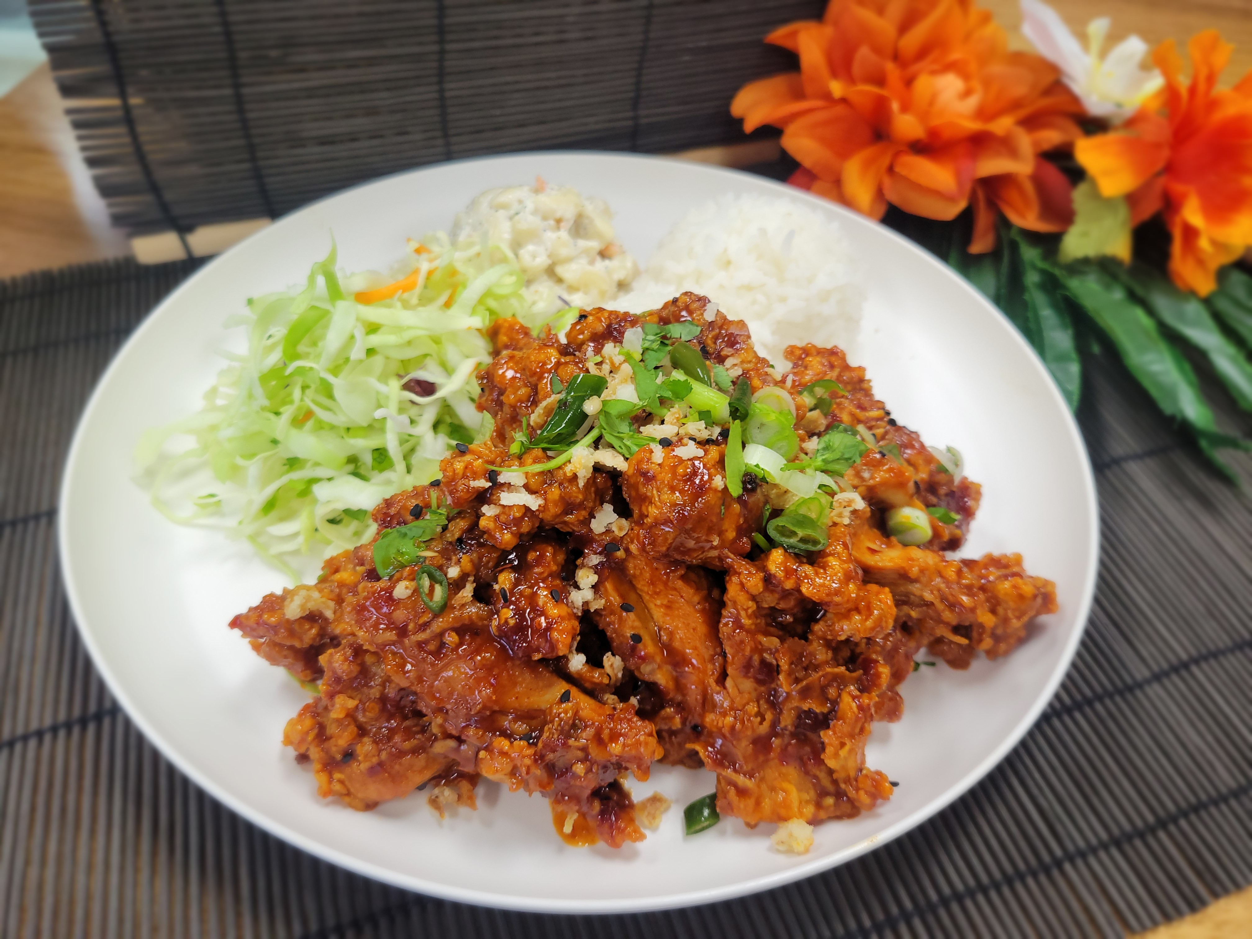 Crispy fried chicken pieces topped with red chili flakes, fried garlic, and fresh green onions in a catering-style box with rice and sides.