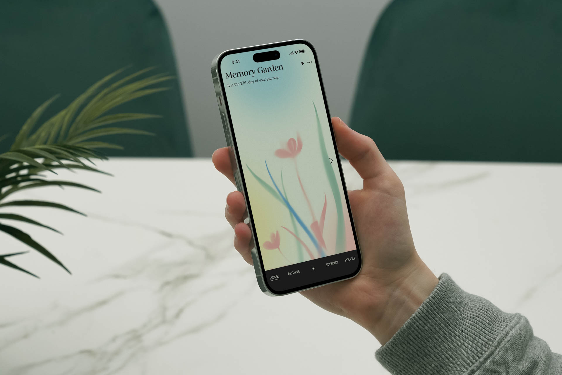 A hand holds a smartphone displaying the app “Memory Garden” on its screen. The screen shows a light blue-green background with pink tulip-like flowers and green stems. The app’s interface includes “Home,” “Activity,” and “Journey” tabs. The phone is held above a white marble surface, with a green plant partially visible to the left and a dark green chair partially visible behind the phone.