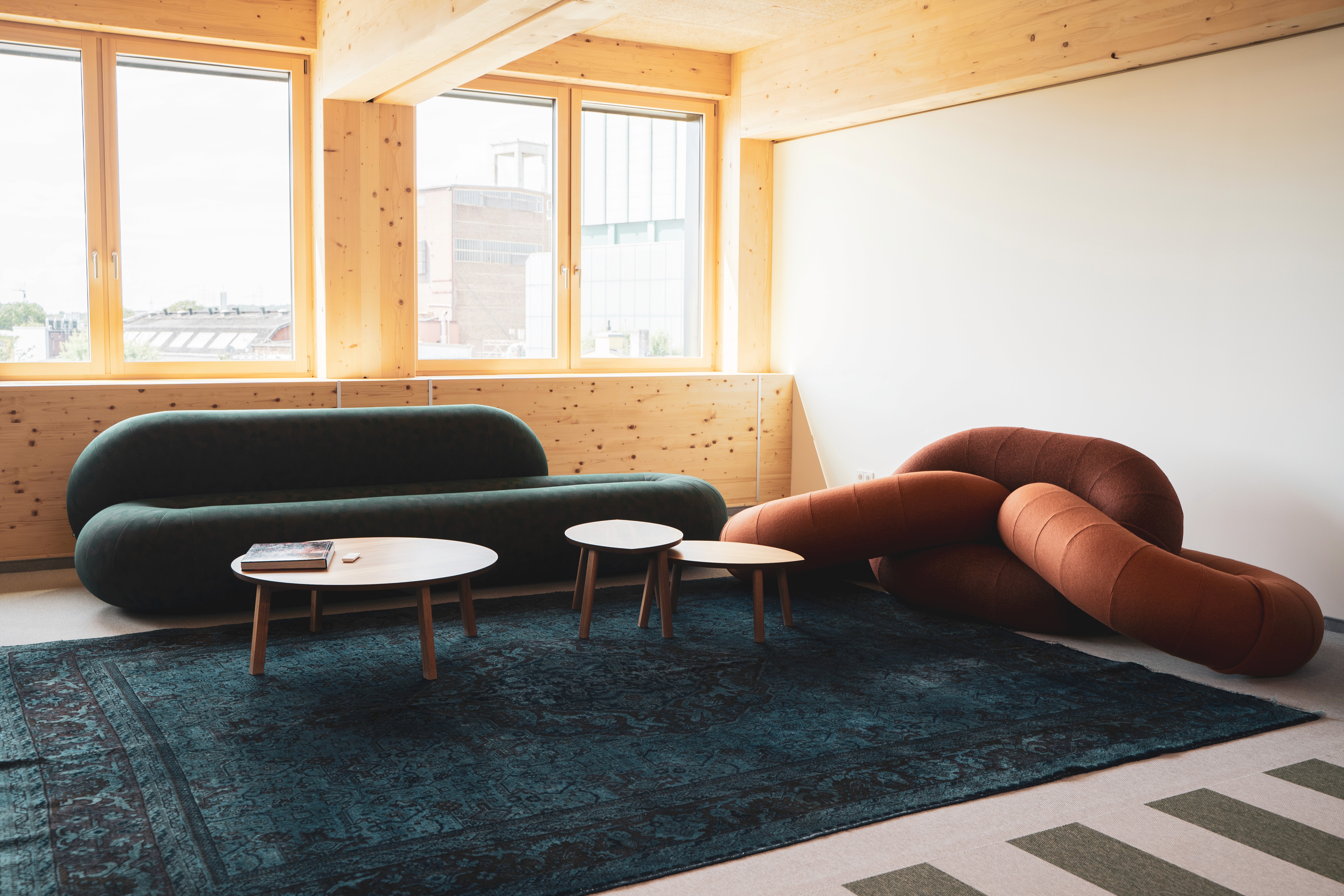 Heller Lounge-Bereich mit moderner Couch, skulpturalem Sitzobjekt und kleinen Tischen auf dunklem Teppich. Raum für kreative Pausen, Austausch und entspanntes Arbeiten.