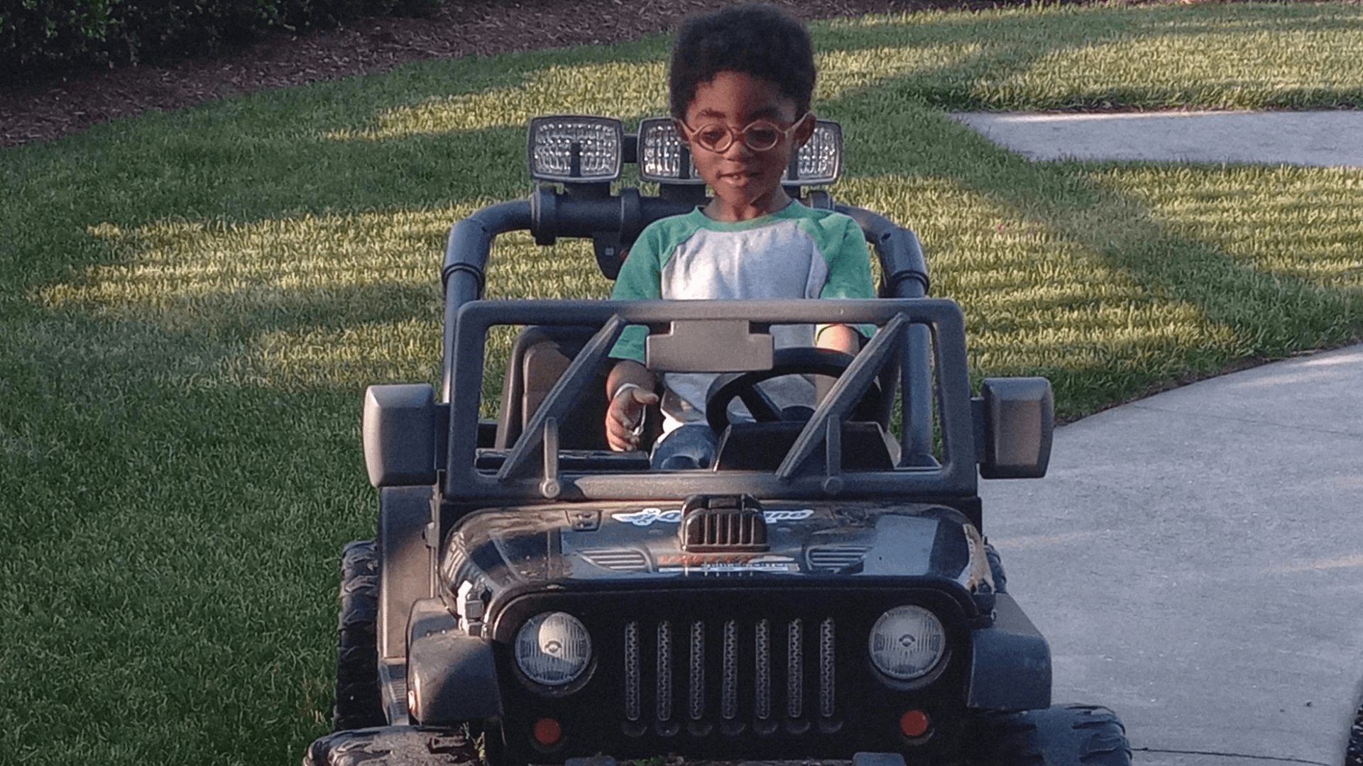 Alt tag: Young child wearing glasses driving a small black ride-on toy Jeep along a sidewalk, smiling and focused outdoors on a sunny day.