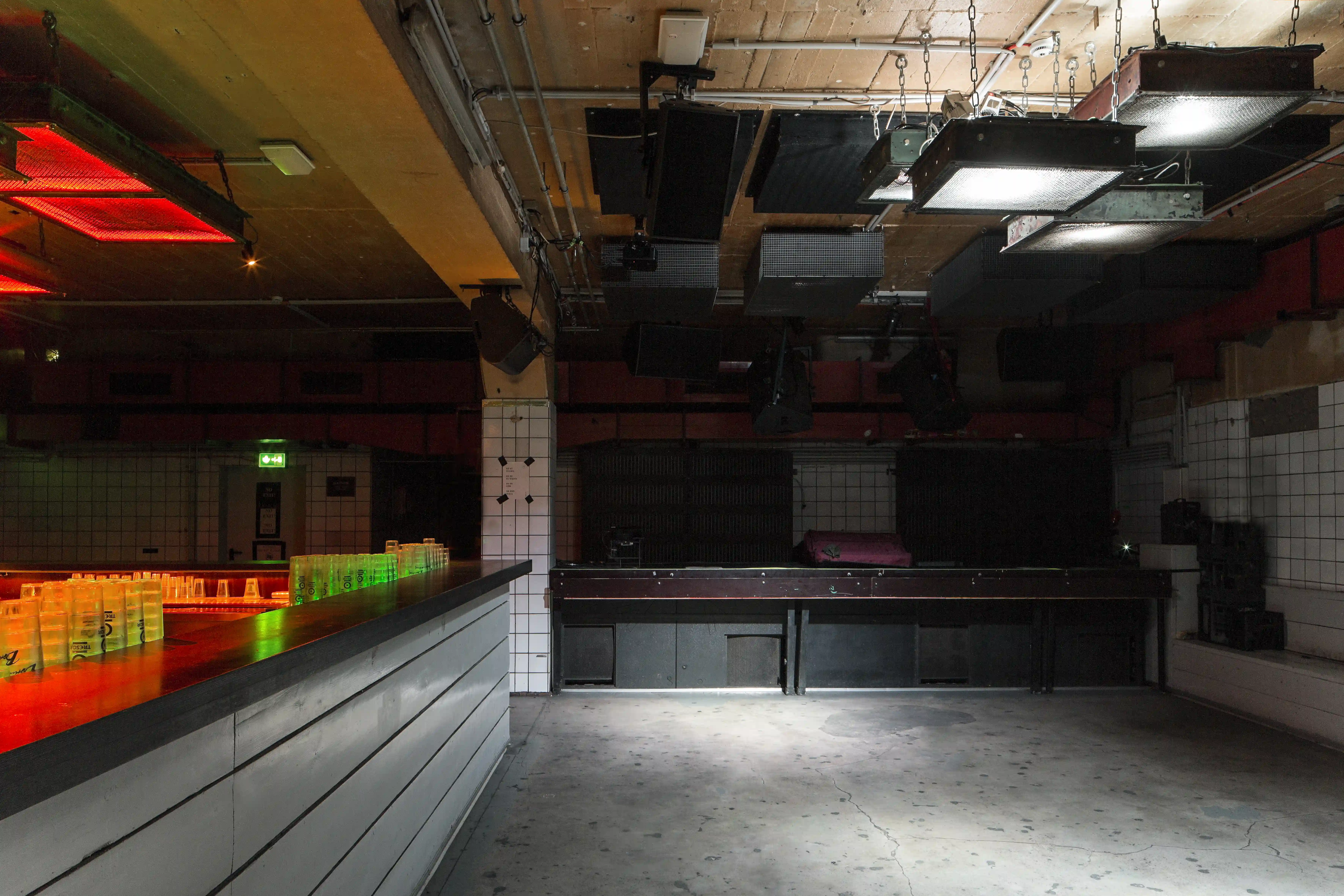 Dark interior of OHM CLUB with the DJ counter and stage area, white tiled wall sections, glowing bottle displays in green and red and black speaker rigs under the ceiling.