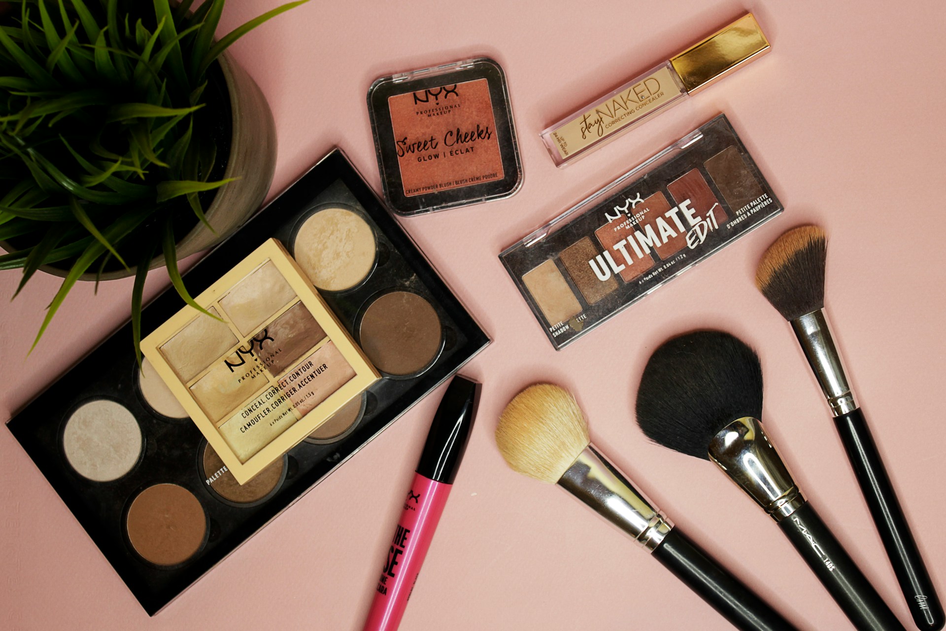 A flat lay photo of several NYX Professional Makeup products and makeup brushes on a light pink background. Products include two face palettes (one large contour/highlight palette and a smaller conceal/correct palette), a blush compact, an Ultimate Edit eyeshadow palette, a tube of lip gloss, and several professional brushes.