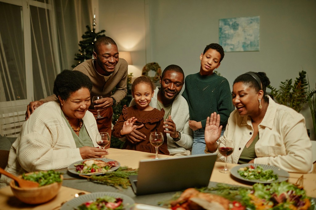 A joyful family gathered around a dining table, smiling and waving at a laptop during a virtual celebration, with a festive meal and Christmas decorations in the background.