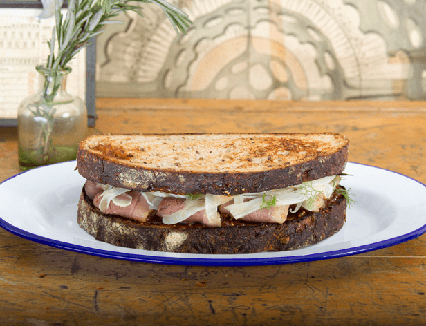 A sandwich made with dark rye bread, layered with pinkish meat and shredded white onions, sits on a white plate with a blue rim. The plate rests on a medium brown wooden table. Behind the plate, a small glass vase containing green stems is visible, and a framed picture or menu is partially seen to the left. A decorative wall panel is visible in the background.