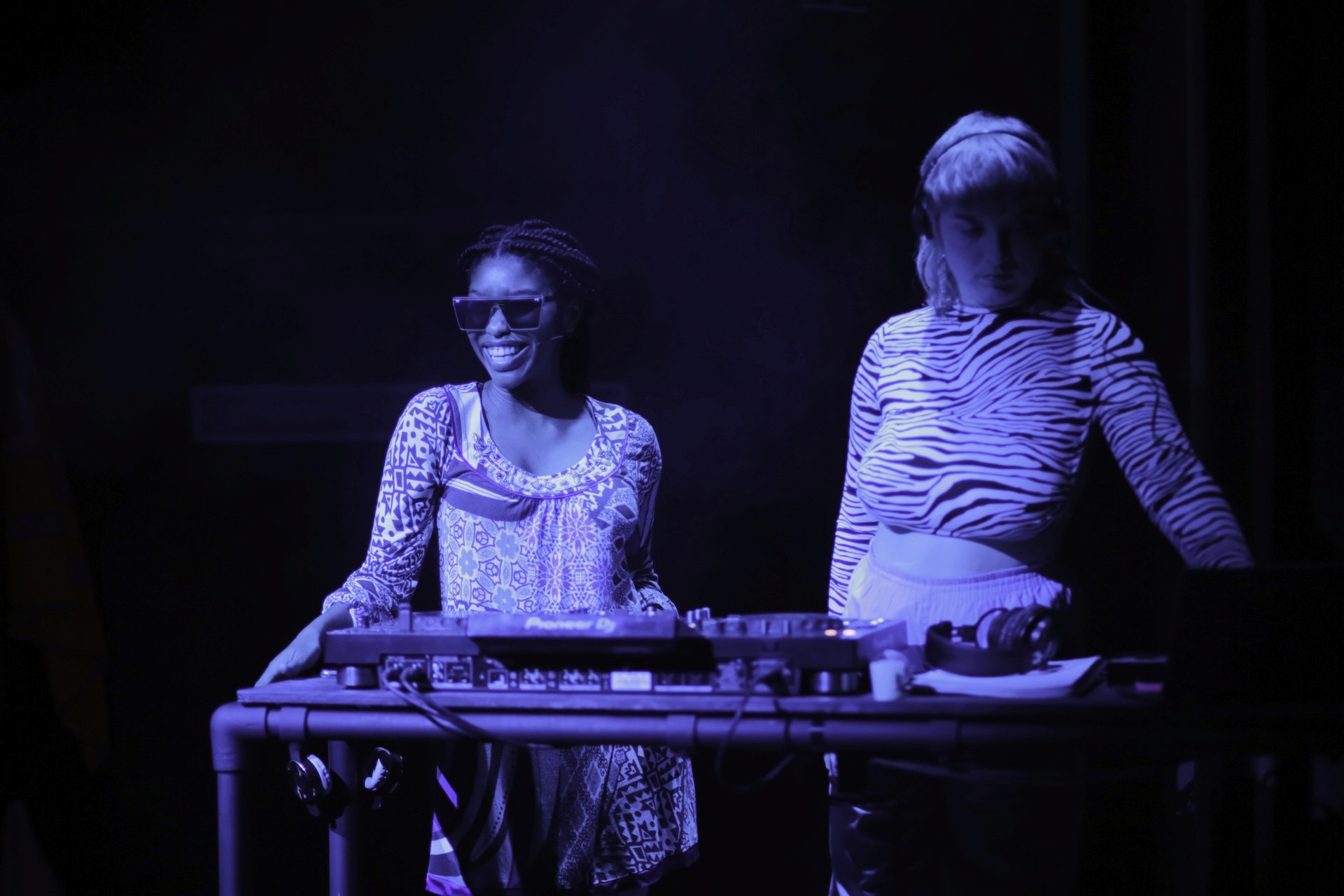 Two performers stand behind a DJ table under deep blue stage lighting. One smiles brightly while wearing large sunglasses and a patterned top, and the other, in a zebra-print long sleeve, focuses on the DJ equipment.
