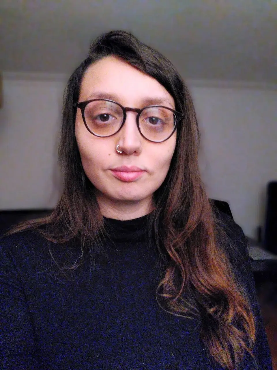 A person with long brown hair and glasses is wearing a black turtleneck, standing indoors against a plain background.