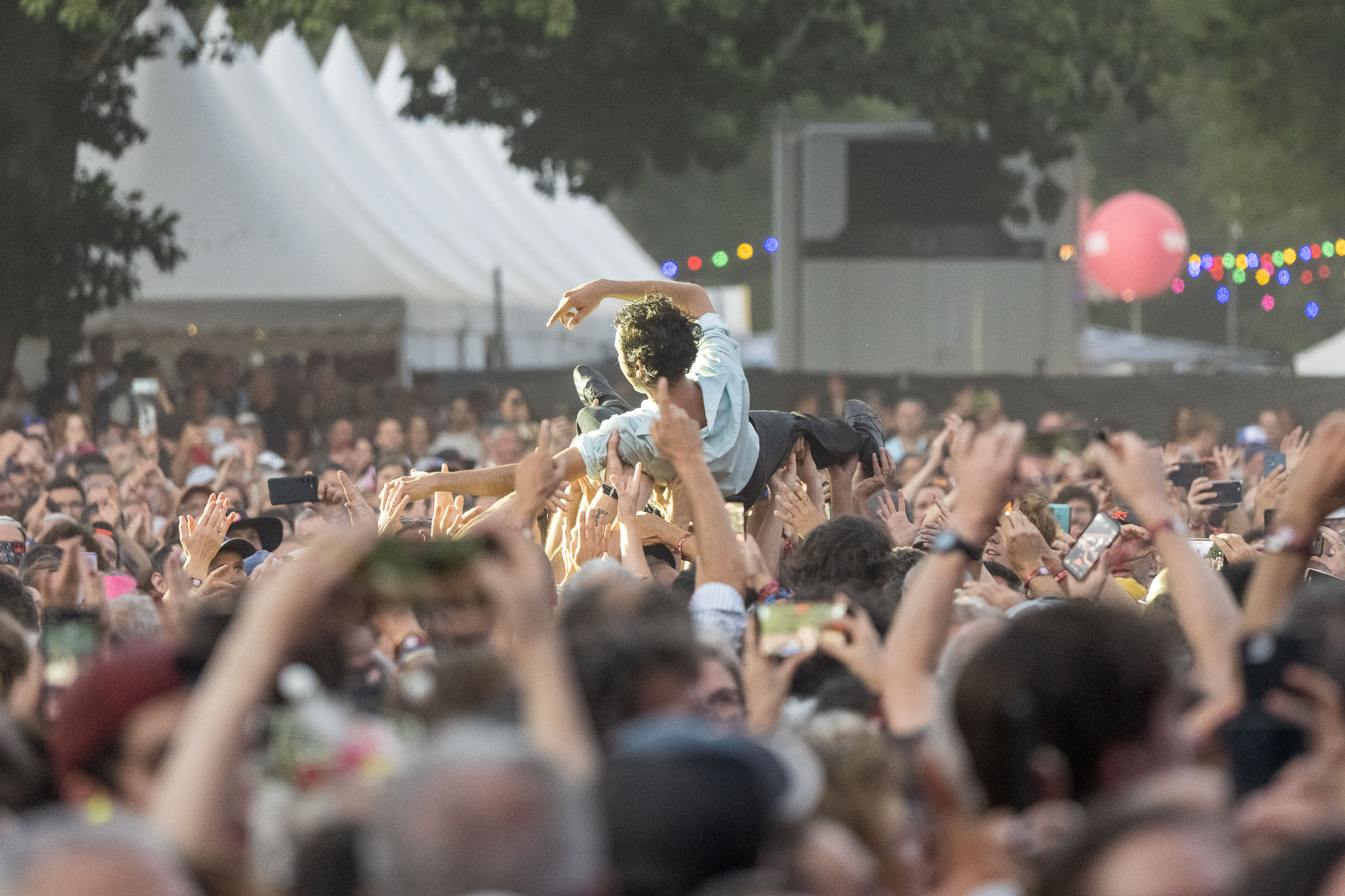 Festivalier porté par la foule, bras en croix, avec une tente blanche et un ballon rose en arrière-plan.