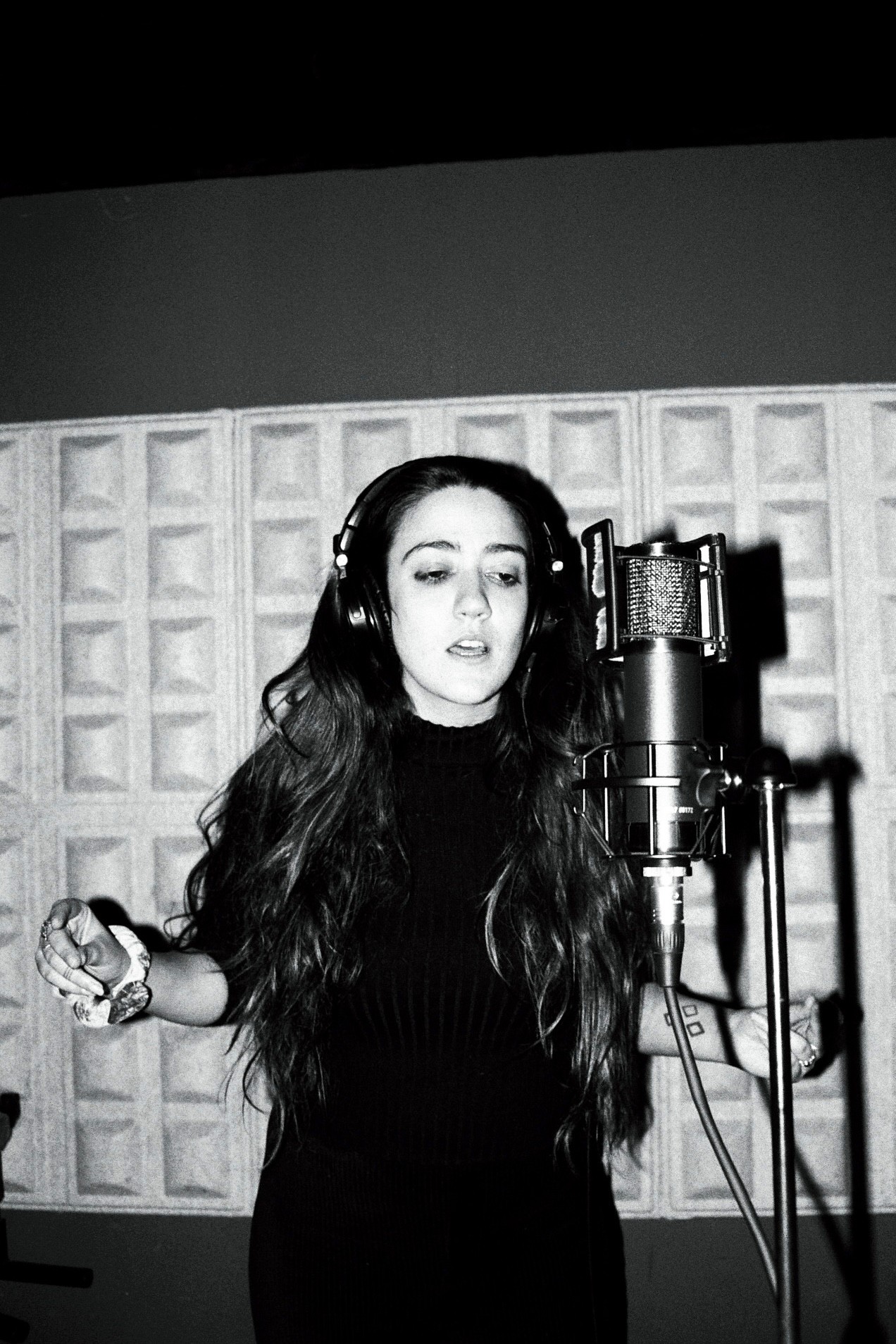 A singer with long hair is posing dramatically in a black-and-white setting, with hands raised and a serious expression.