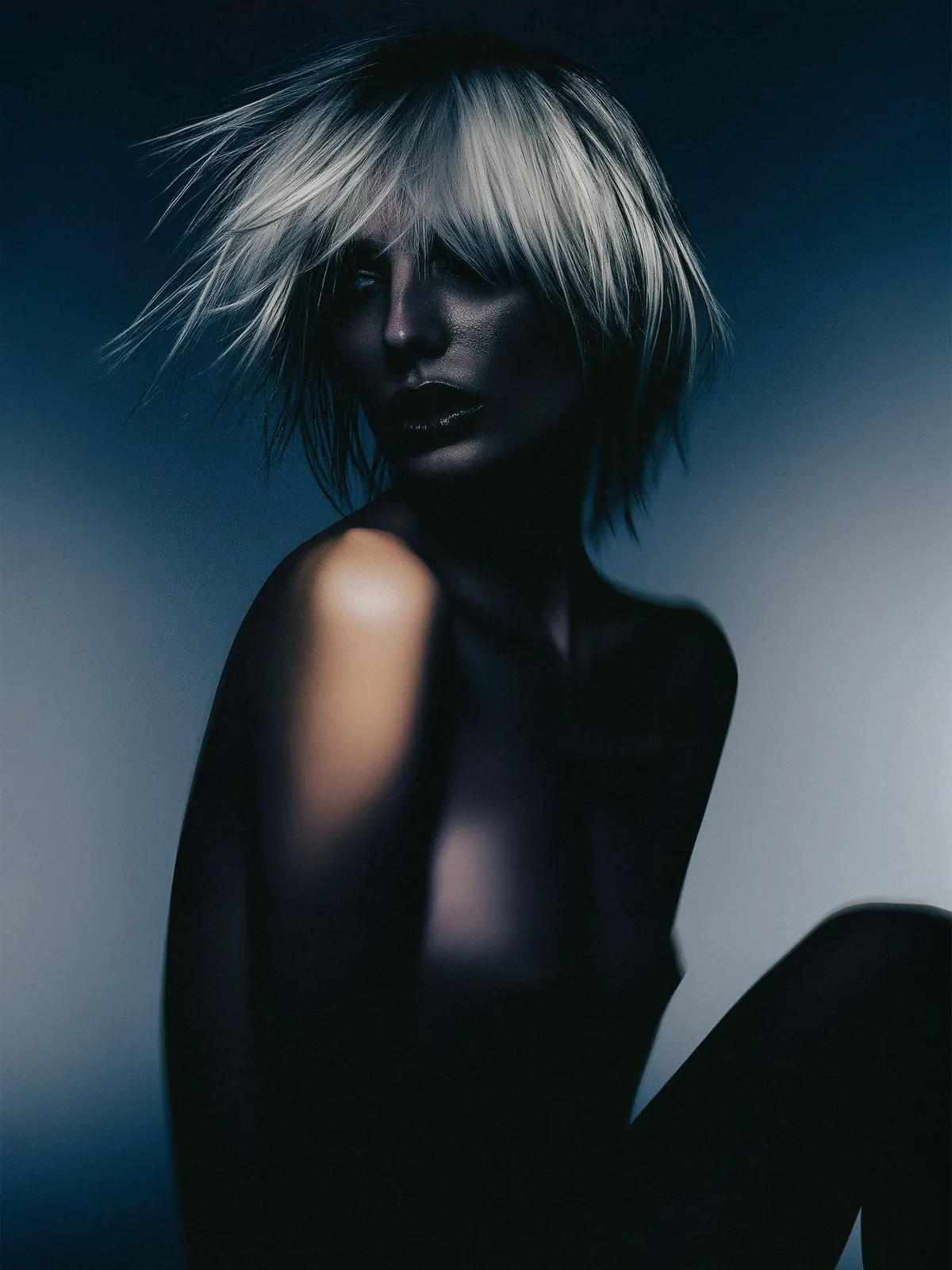 Fashion portrait with a tousled blonde bob and a bare shoulder in cool low light.