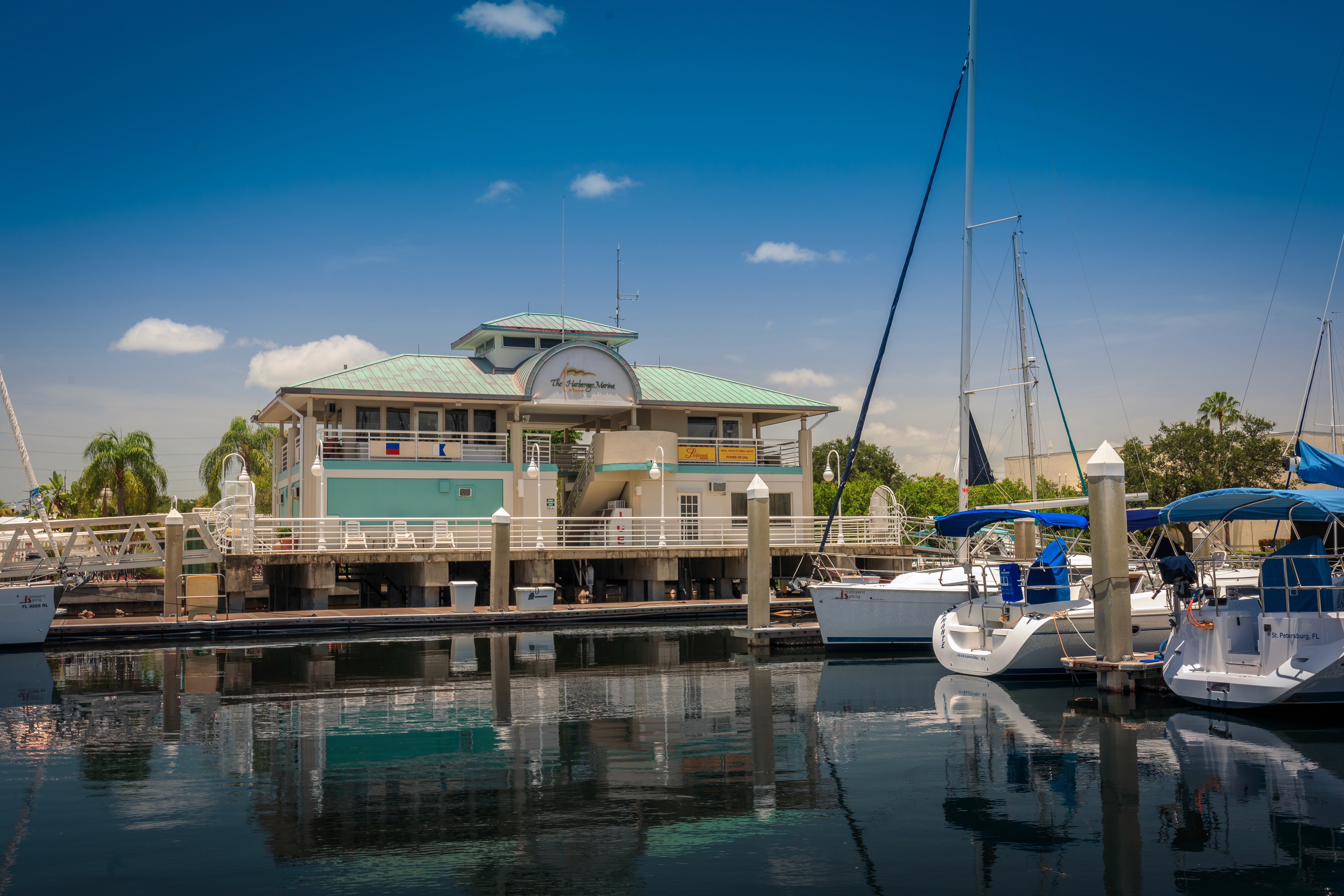 A Harborage marina at St Pete, FL scene featuring a turquoise-roofed nautical building surrounded by several sailboats, with calm waters reflecting the vibrant blue sky and fluffy clouds, creating a tranquil seaside atmosphere.