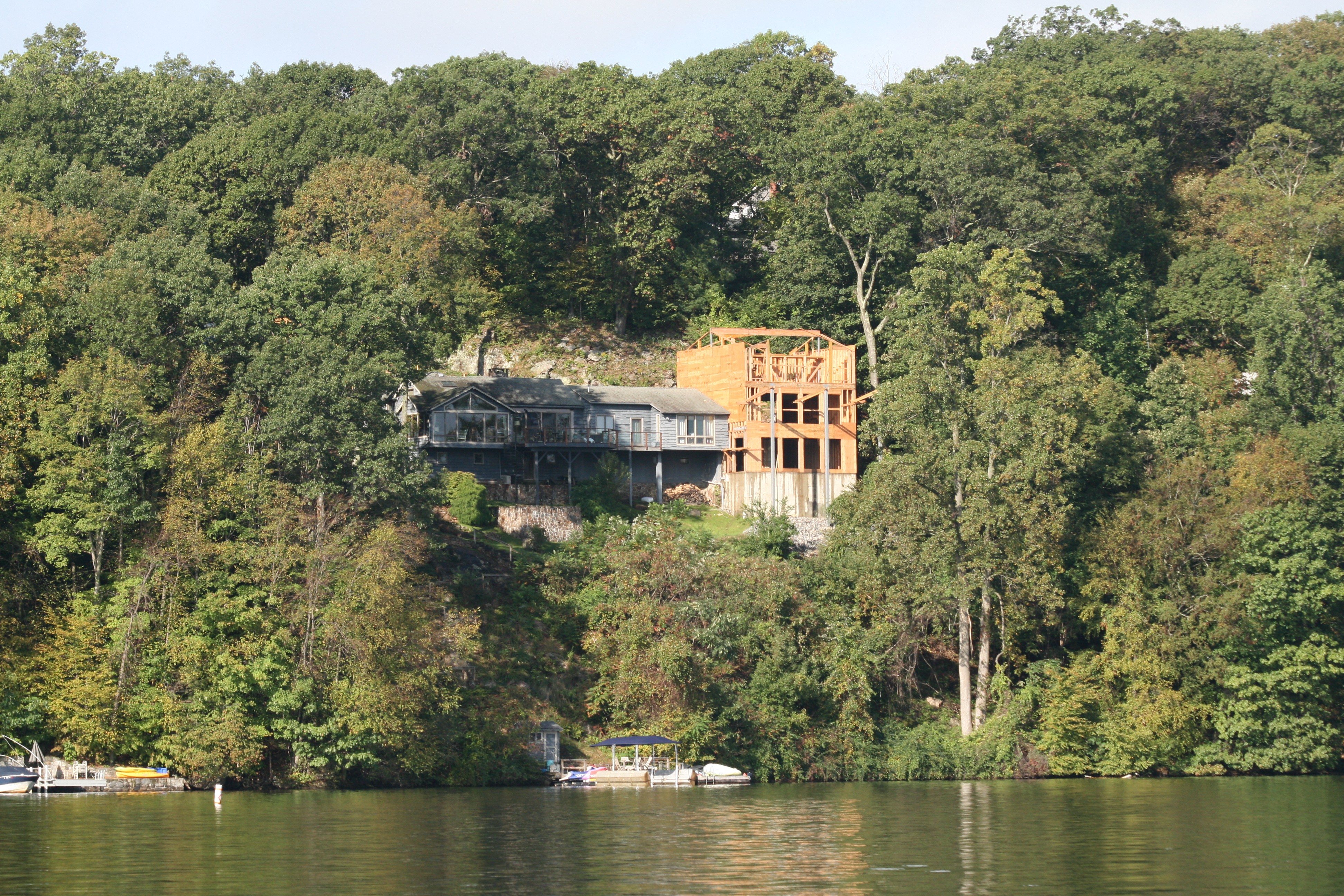 Lakefront home with new multi-story wood-framed addition under construction beside existing house on wooded hillside.