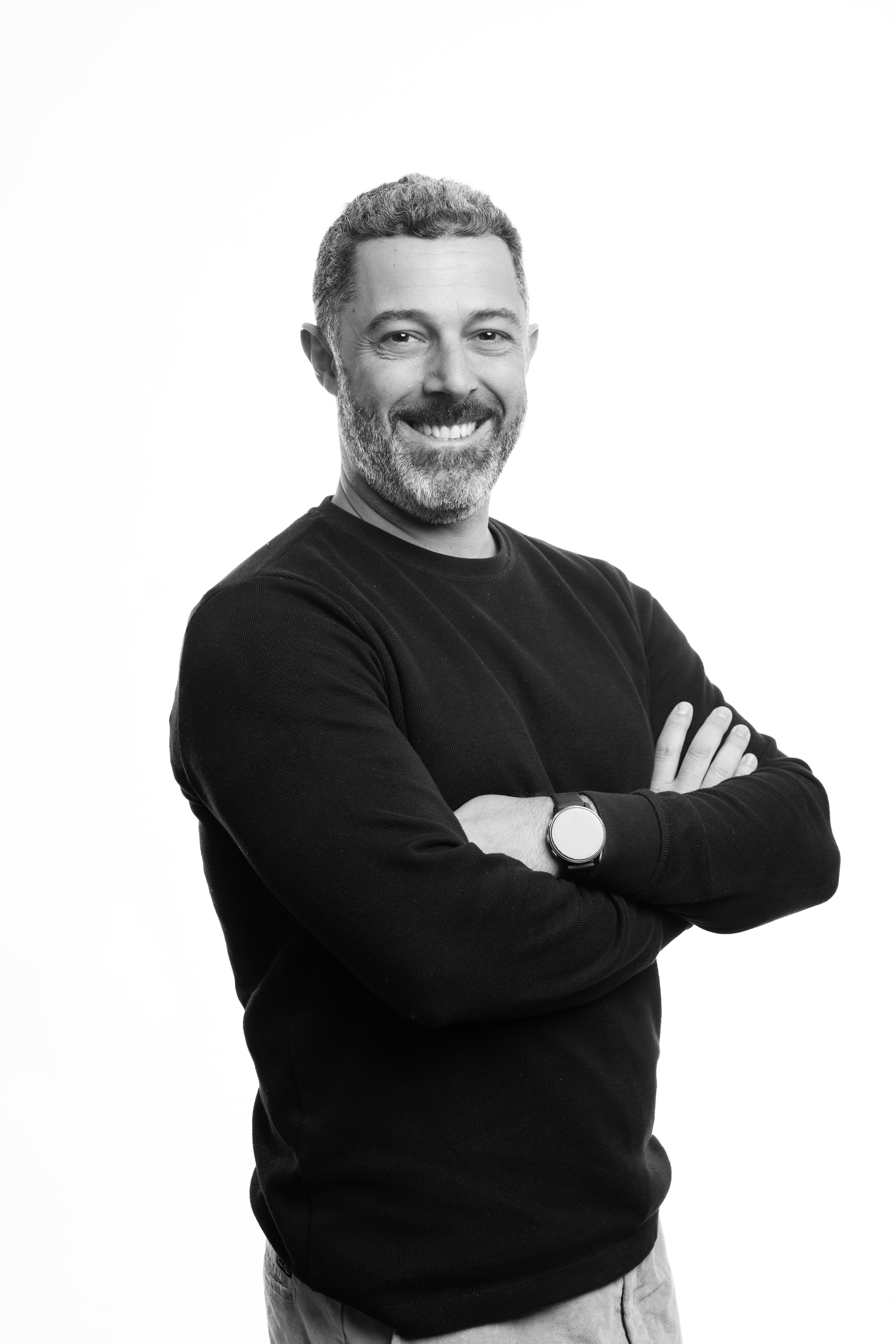 A man with a beard and short curly hair smiles confidently, crossing his arms. He wears a dark sweater and a watch, set against a plain white background.