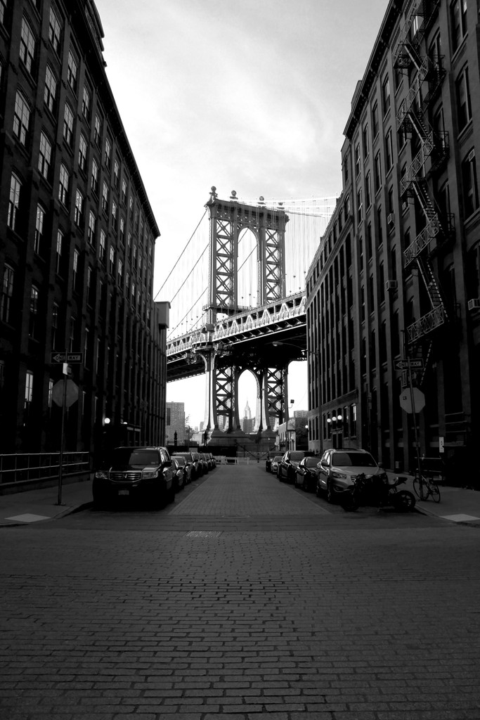 black white image of a suburban street with builidngs to each side and the brooklyn bridge revield in the middle of the shot.