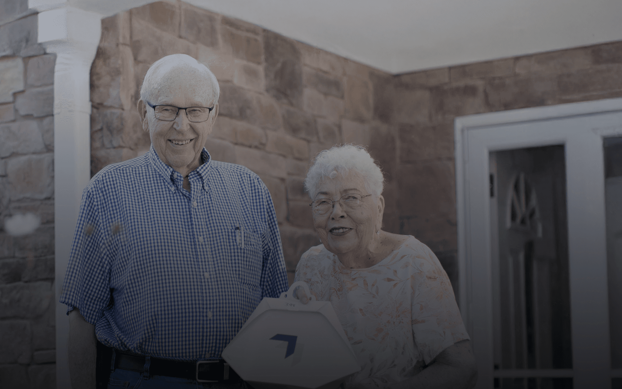 Two elderly people stand outside a brick house, smiling and holding a white object shaped like a house with a blue logo, against a backdrop of stone walls and a partially visible door.