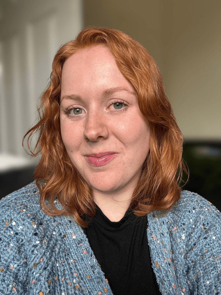 Eve with shoulder-length red hair smiling at the camera indoors, wearing a blue knitted cardigan over a black top.