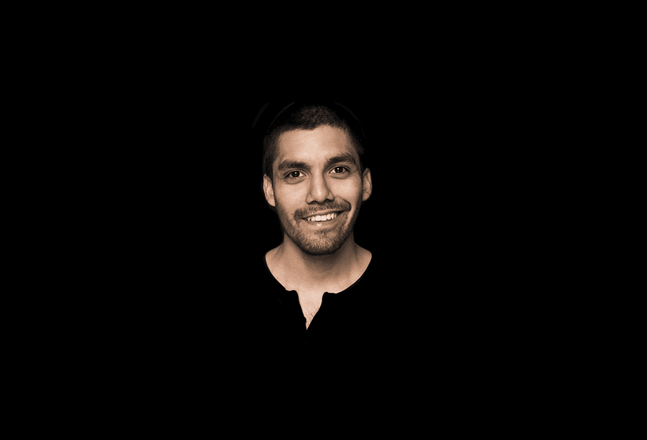 A man with short dark hair and a beard is smiling at the camera against a solid black background. He is wearing a dark-colored shirt with a collar. The lighting highlights his face and upper body.