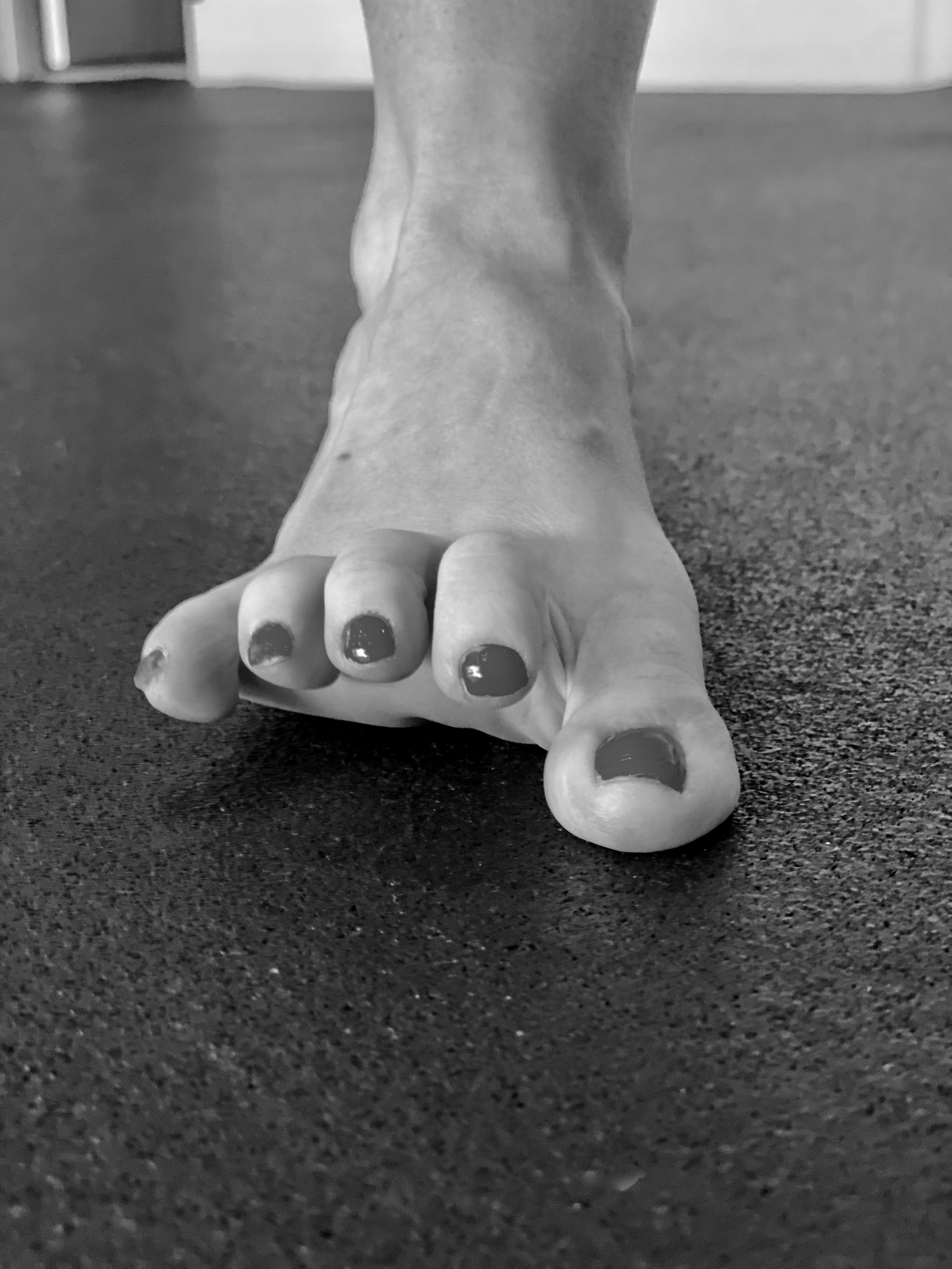 A black and white image shows a person's foot with painted toenails stretching out on a textured, dark surface, suggesting a focus on foot flexibility or stretching exercises.