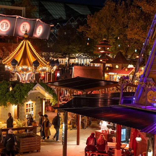 En natfestival med farverige lys, en karrusel, mennesker der går rundt, og forskellige boder og dekorationer.
