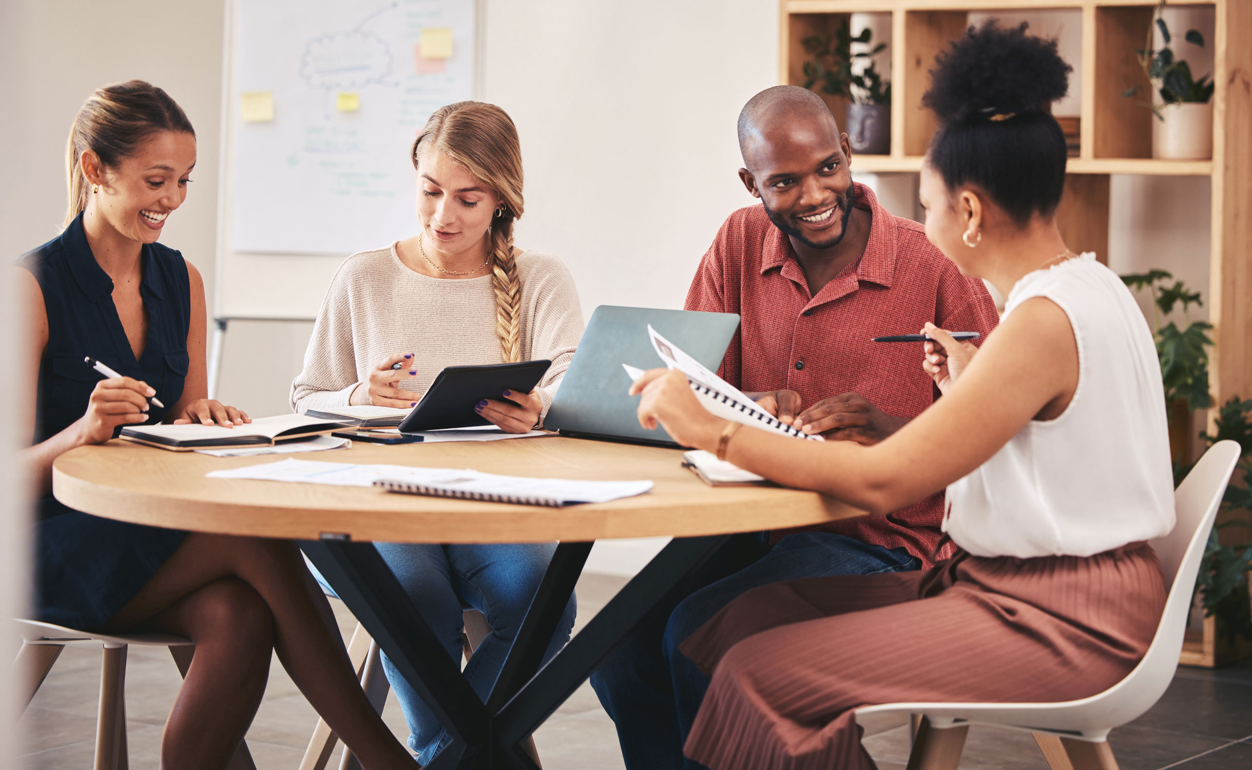 Czworo uśmiechniętych specjalistów podczas kreatywnego spotkania przy okrągłym stole omawiających strategię reklamową z użyciem laptopa i tabletu