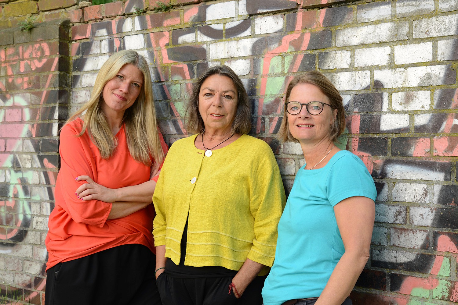 Die drei Gesellschafterinnen der school of facilitating stehen vor einer Wand mit Graffiti.