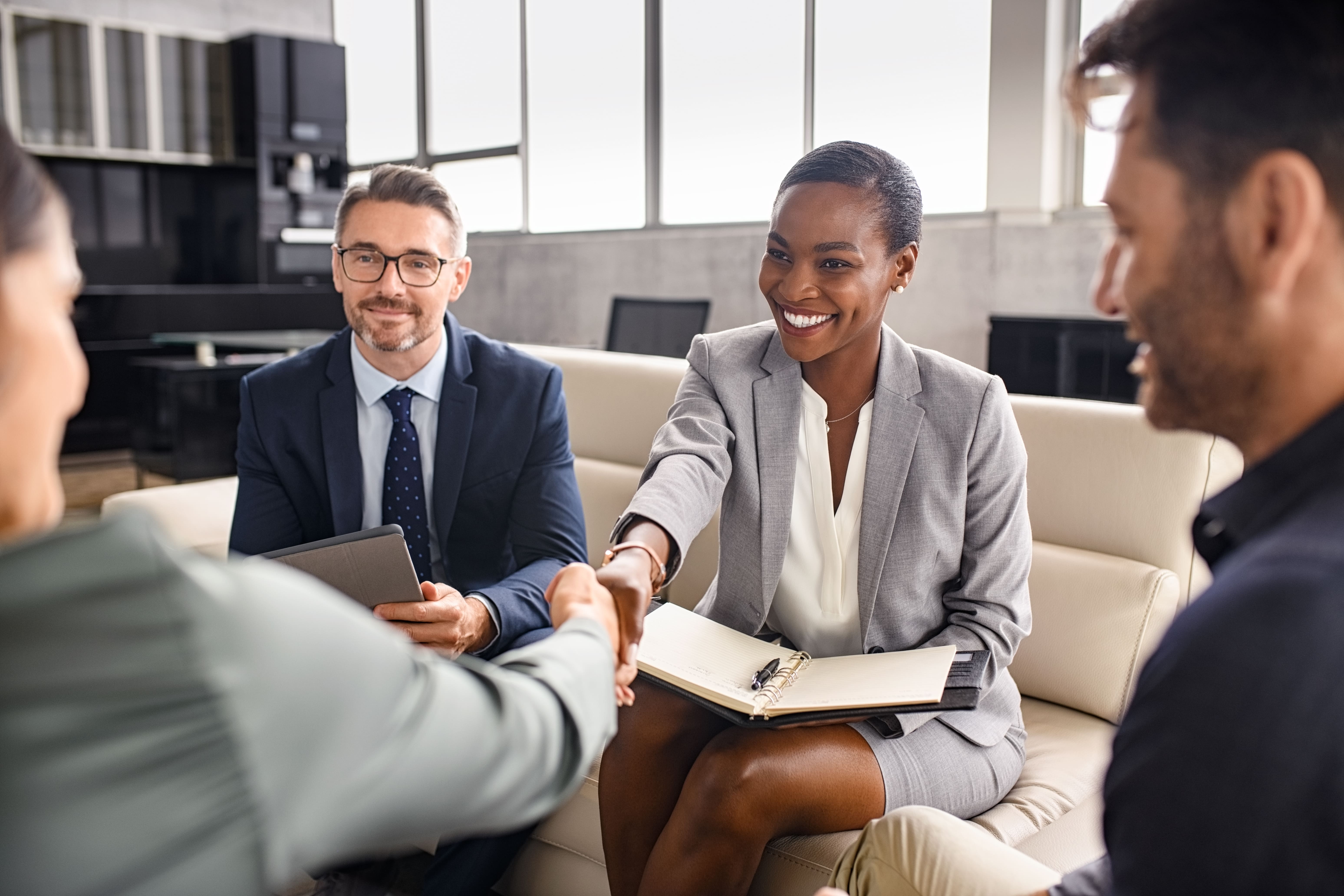 Imagem de uma reunião de negócios onde quatro pessoas estão envolvidas. Uma mulher sorridente de terno cinza aperta a mão de outra pessoa, sugerindo o fechamento de um acordo ou uma apresentação cordial. Ao lado dela, um homem de óculos em um terno escuro observa a interação com um leve sorriso, segurando um tablet. Outras duas pessoas, parcialmente desfocadas, completam o grupo, criando uma atmosfera profissional e positiva, enfatizando a colaboração e o sucesso no ambiente corporativo.