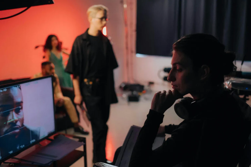 A person watches a monitor showing a man's face. Others stand on a film set in the red-lit background.